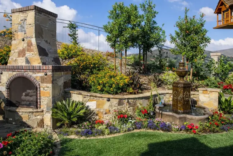 Stone fireplace and fountain surrounded by colorful flowers in a landscaped garden.