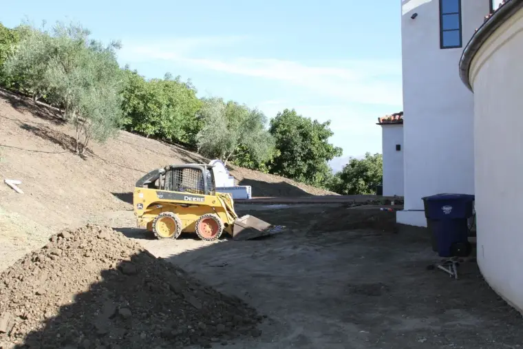 Construction site with a yellow skid steer loader and freshly dug earth beside a residential building.