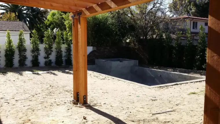 Backyard construction site with a concrete pool under a wooden pergola and lush greenery.