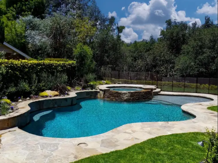 Elegant backyard pool with a spa, surrounded by lush greenery and a stone patio.