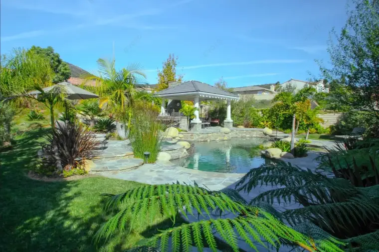 Lush backyard oasis with a pool, gazebo, palm trees, and landscaped greenery.