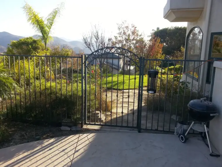 Metal gate opening to a scenic garden with mountains in the background and grilling equipment nearby.