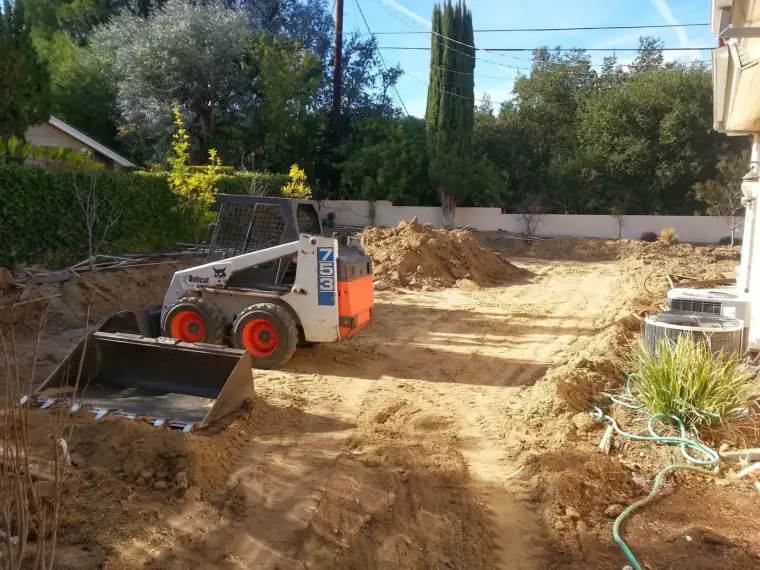 Bobcat skid steer loader on a dirt site with trees and a house in the background.