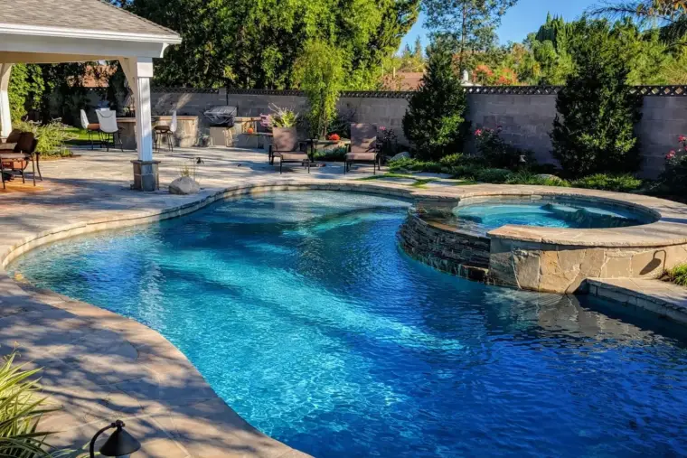 Luxury backyard swimming pool with stone accents, lounge chairs, and lush landscaping.