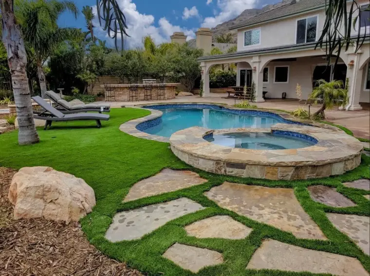 Backyard oasis with a pool, spa, stone pathway, and lounging chairs surrounded by greenery.