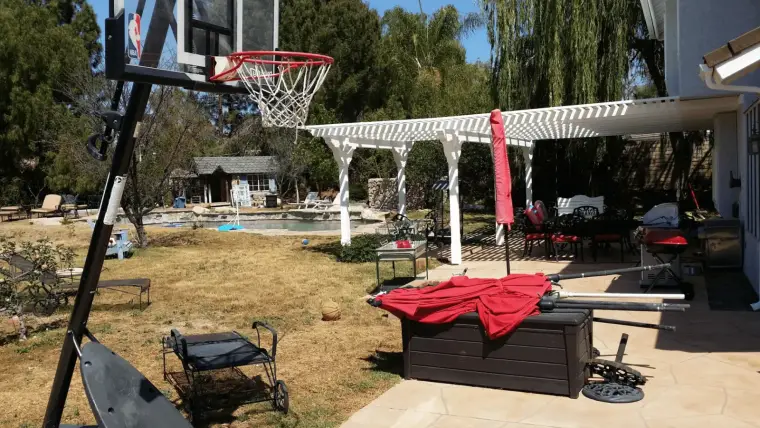 Backyard scene featuring a basketball hoop, pool, and patio furniture under a pergola.
