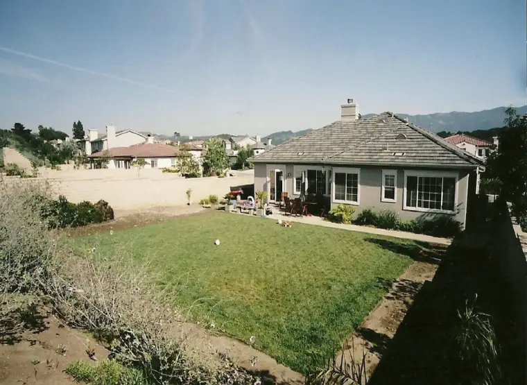 House with backyard lawn and patio furniture under clear blue sky. Mountains in background.