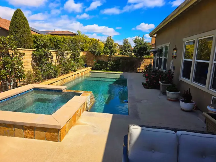 Modern backyard pool with spa, surrounded by lush greenery and a blue sky.