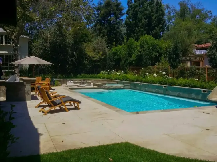 Backyard pool area with sun loungers, umbrella, and lush greenery in a sunny setting.