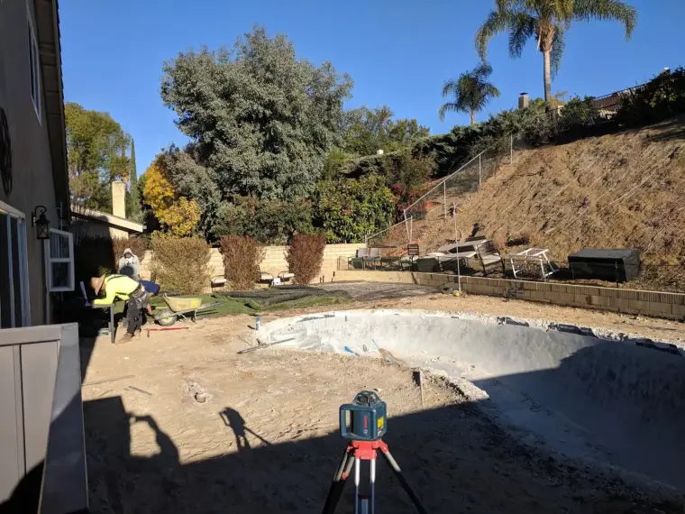 Pool construction site with workers, surveying equipment, and landscaped yard.