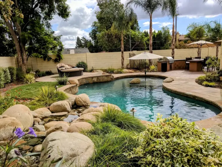 Lush backyard pool with rock features, palm trees, and patio furniture under a cloudy sky.