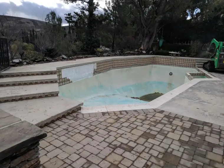 Empty swimming pool with cracked tiles, surrounded by stone patio and lush landscaping.