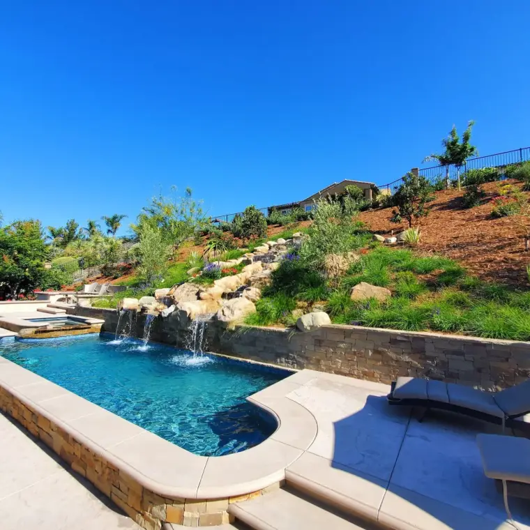 Beautiful backyard pool with waterfalls, stone features, and landscaped hills under a clear blue sky.