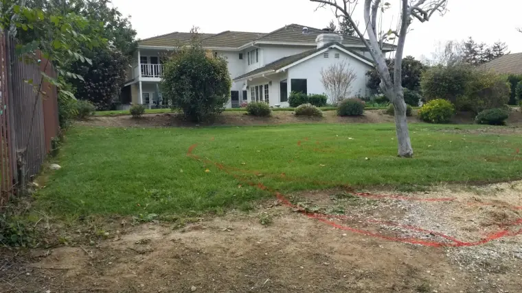 Lawn with marked area and residential home amid trees and shrubs in background.