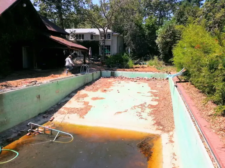 Abandoned swimming pool undergoing renovation with overgrown landscape and nearby house.
