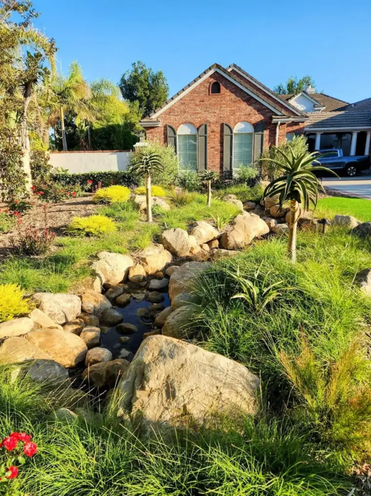 Lush garden with a stone creek and a brick house in the background under a clear blue sky.