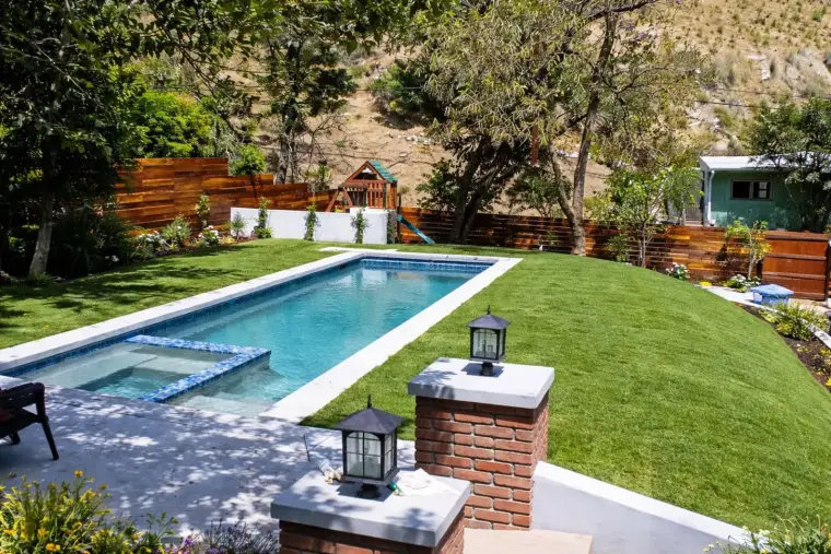 Modern backyard pool with landscaping, grassy area, and playset in a sunny outdoor setting.