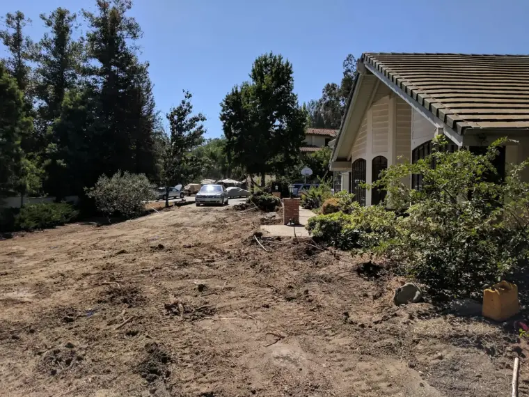 Residential landscape with cleared dirt area, house, trees, and parked cars under blue sky.