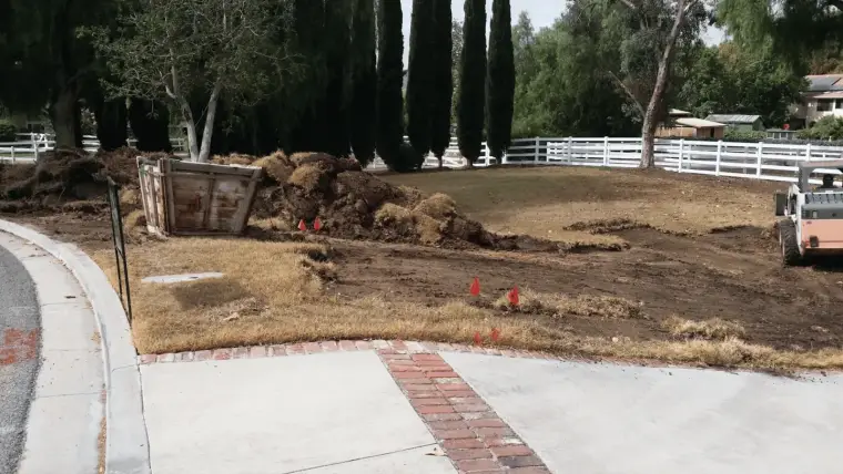 Construction site showing dirt piles, a dumpster, and heavy machinery near a residential area.