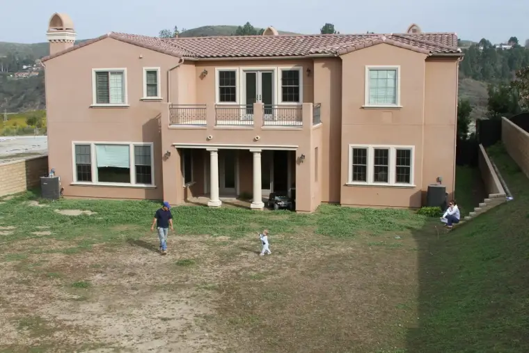 Two people walk in front of a large two-story home with a backyard and hills in the background.