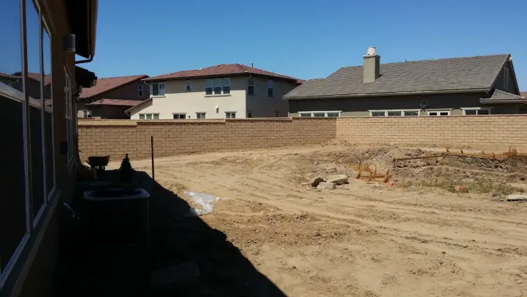 Empty, cleared lot with dirt surface and surrounding houses under a clear blue sky.