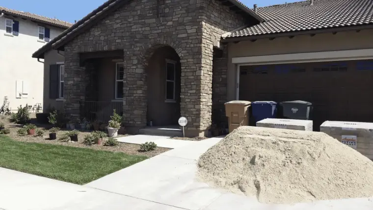 Residential landscaping with a stone house, a sand pile, and recycling bins outside.
