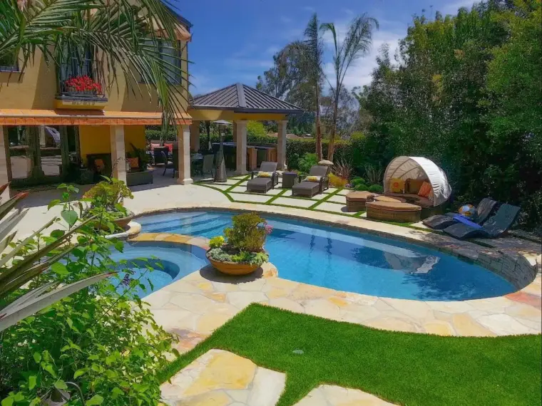 Lush backyard with a swimming pool, lounge chairs, and tropical plants under a clear blue sky.