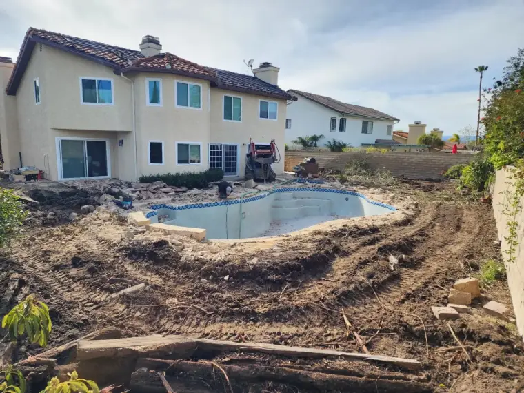 Construction site with an empty pool and two houses in the background, showcasing landscaping work.