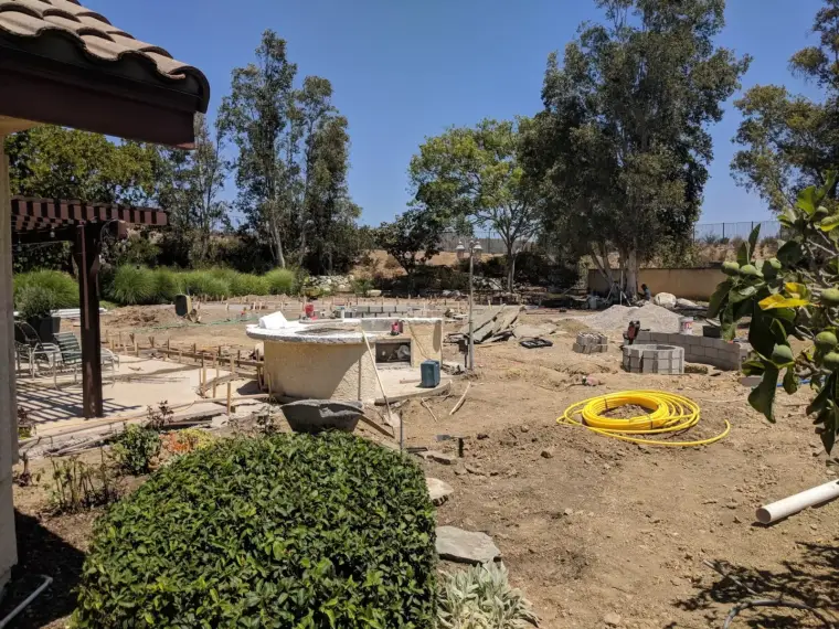 Construction site in a landscaped yard with scattered materials and trees in the background.