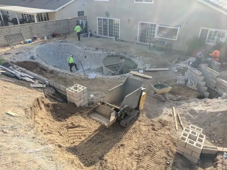 Construction workers building a swimming pool in a residential backyard. Heavy machinery nearby.