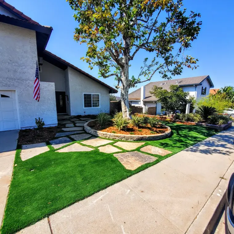 Lawn with artificial grass and stone pathway, bordered by plants and trees near a house.