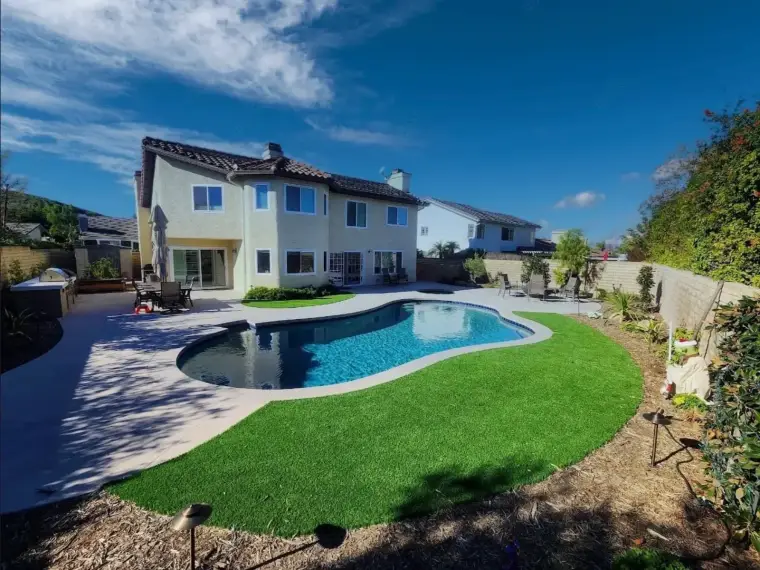Modern backyard with a swimming pool, lush green lawn, and a two-story house under a blue sky.
