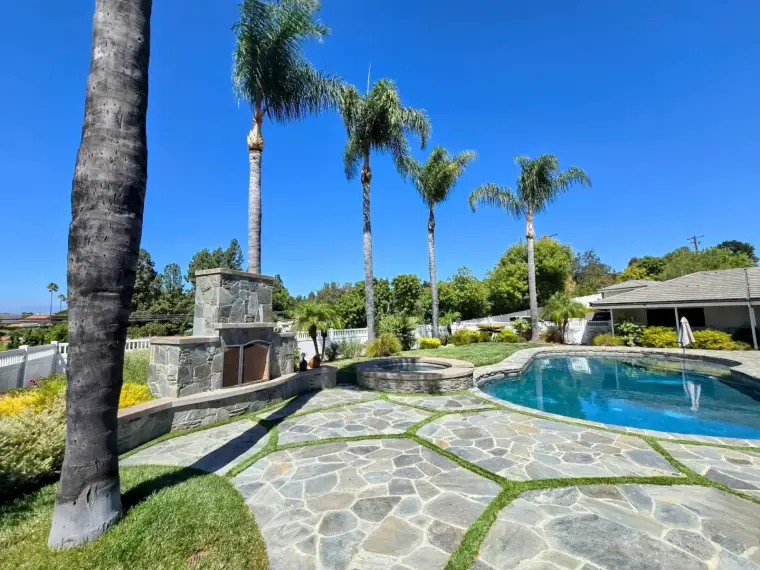 Luxury backyard with a stone patio, swimming pool, palm trees, and outdoor fireplace.