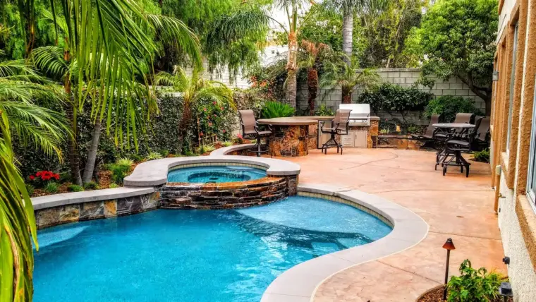 Luxury backyard patio with a pool, spa, and barbecue area surrounded by lush greenery.