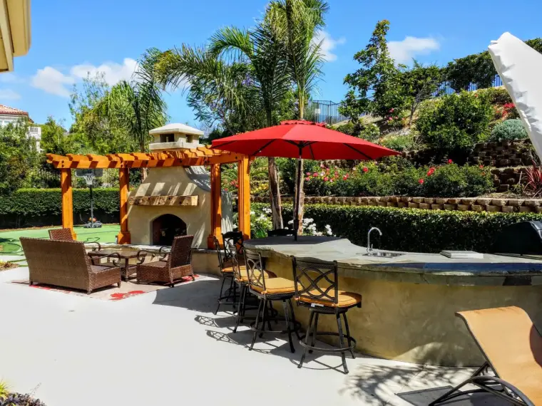 Outdoor patio featuring a stone bar, red umbrella, and lounge seating surrounded by lush greenery.
