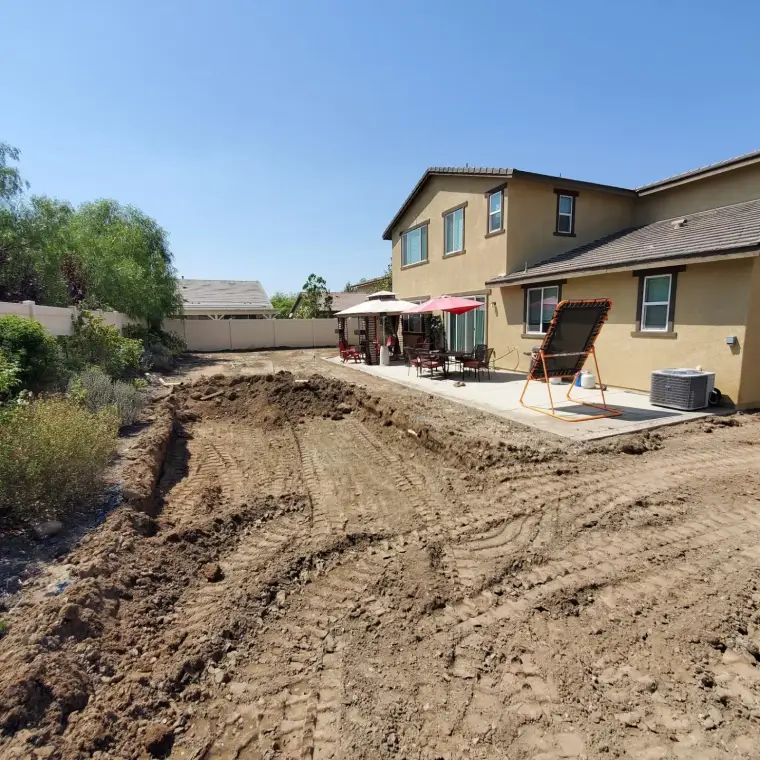 Backyard with freshly dug ground, patio area, tables, chairs, and a house in clear sunny weather.