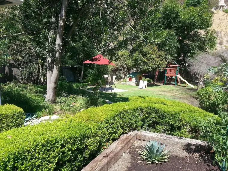 Sunny backyard with green hedges, a red umbrella, and a playground set for children.