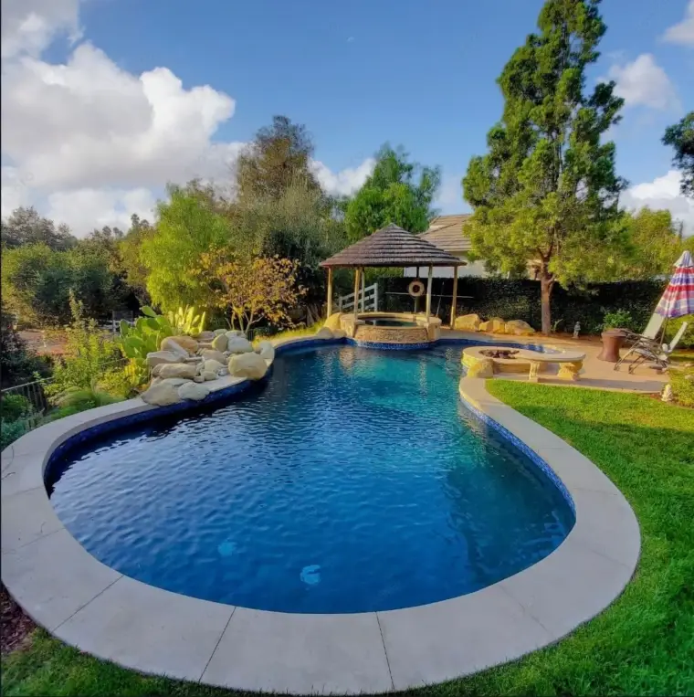 Serene backyard with a curved pool, gazebo, and lush greenery under a partly cloudy sky.