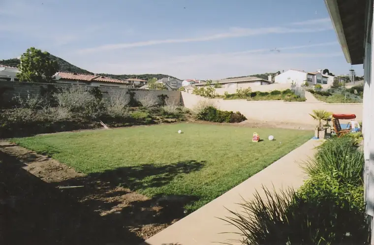 Child playing in a green backyard with a wall, houses, and mountains in the background.