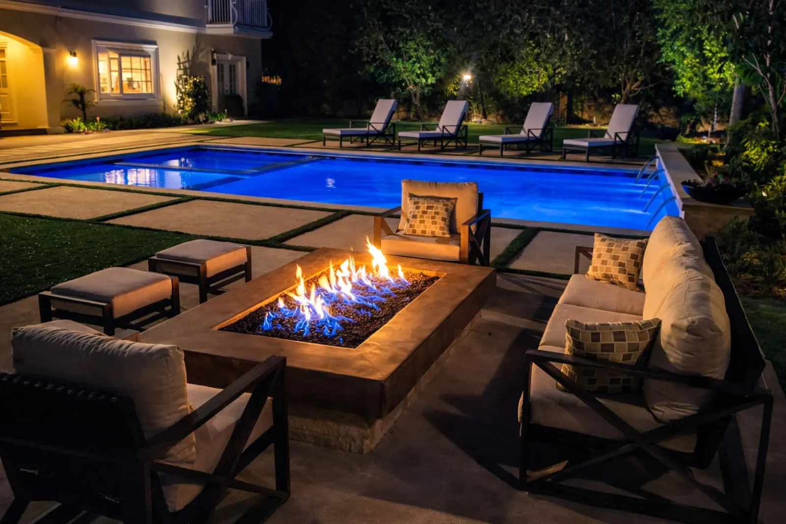 Elegant outdoor patio with fire pit, pool, and lounge chairs at night.
