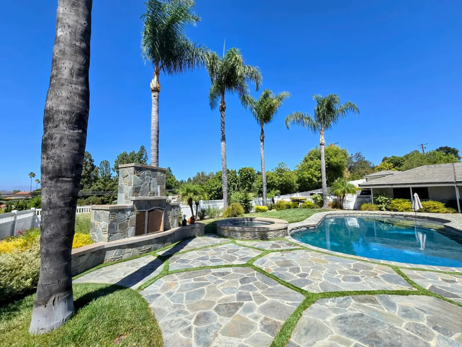 Luxury backyard with a stone patio, swimming pool, palm trees, and outdoor fireplace.