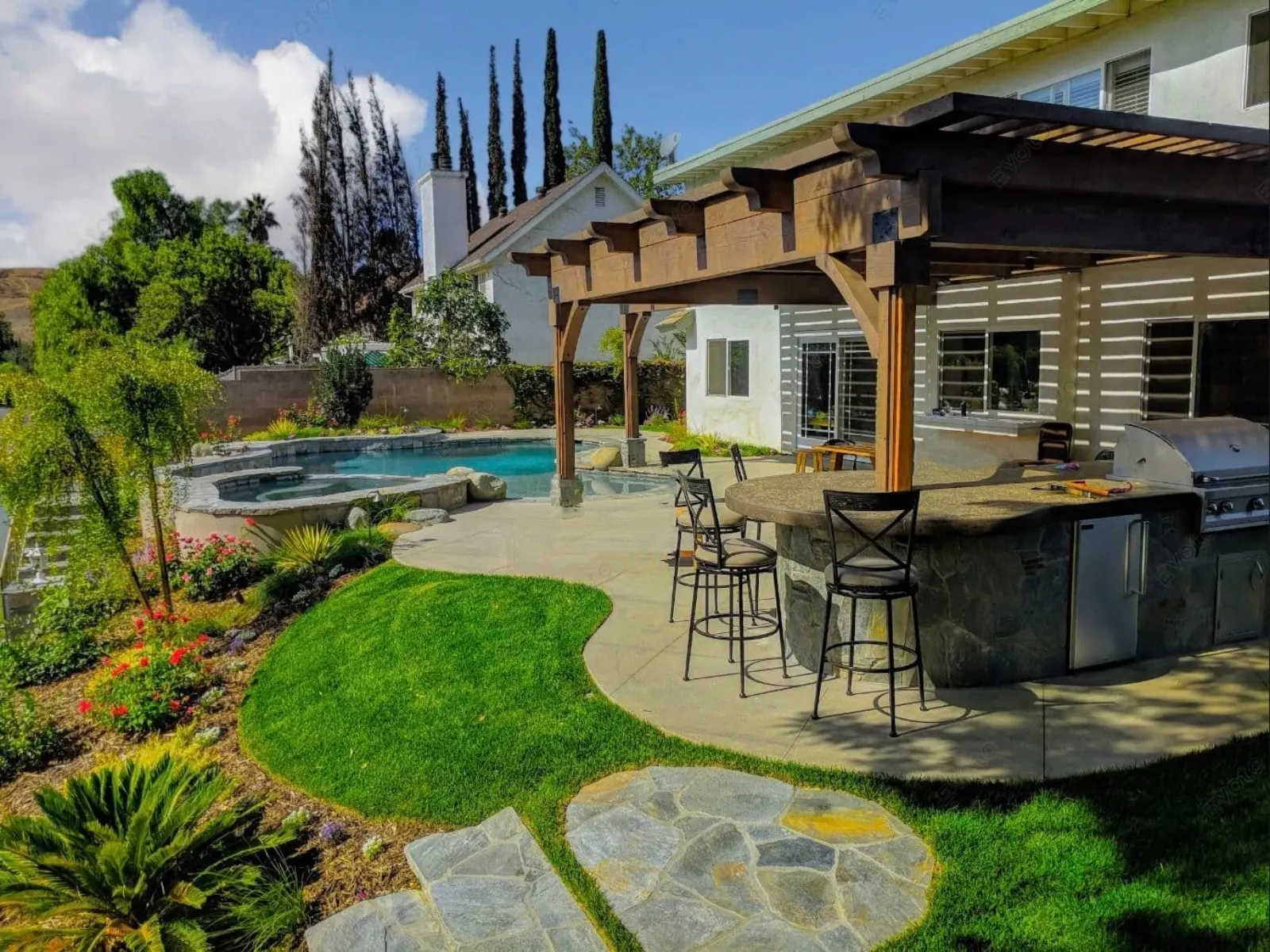 Backyard patio with barbecue, bar seating, pool, and lush landscaping under a pergola.